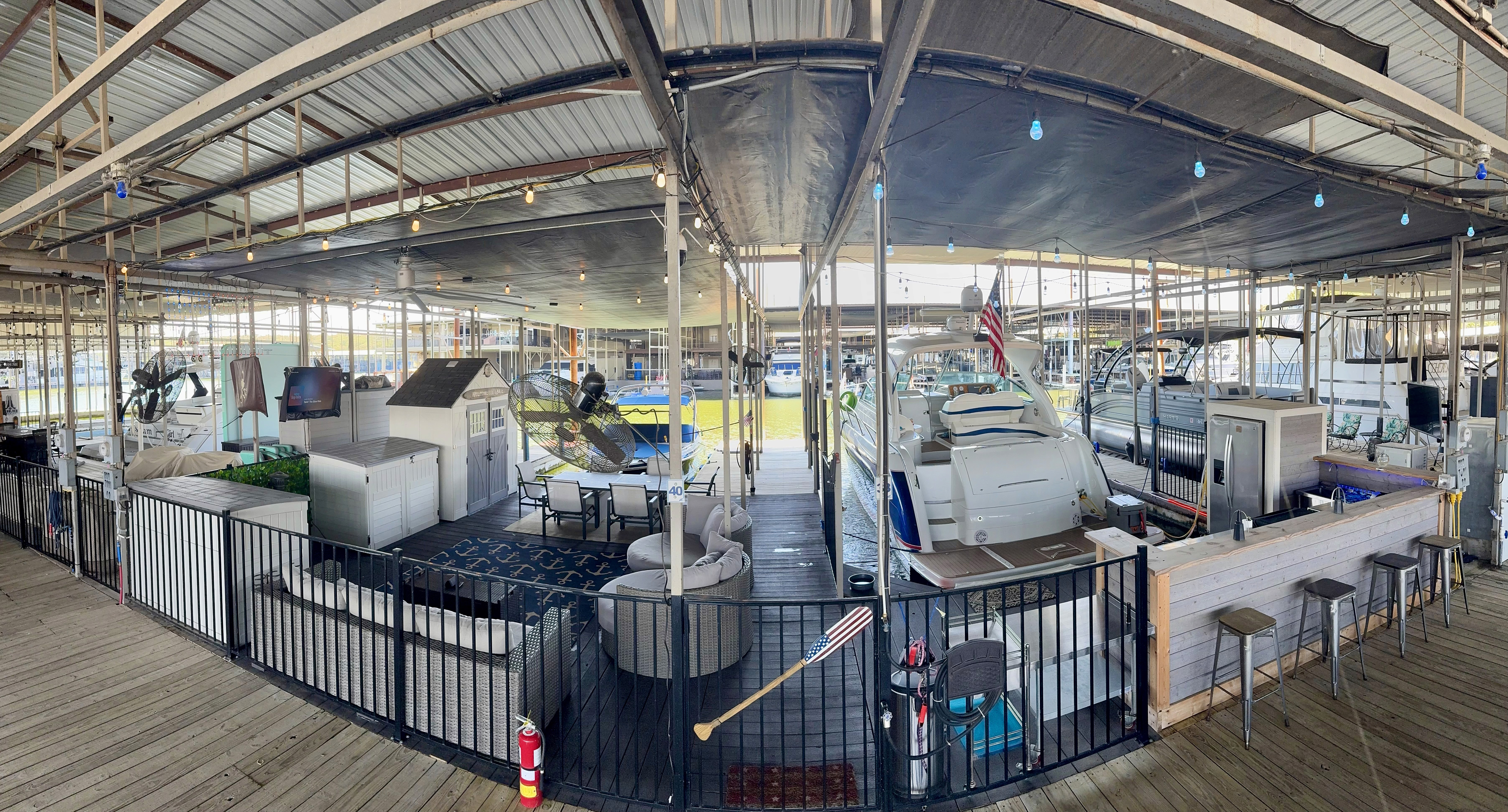 Panoramic view of a spacious marina with multiple docked boats covered by a metal roof, featuring cozy seating areas, tables, and nautical decor, including an American flag and a golf club, against a backdrop of bright lights and water.