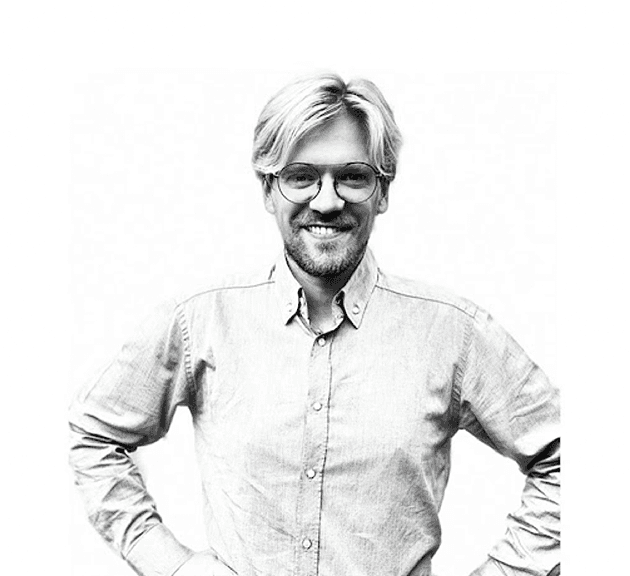 A focused woman with long red hair is seated at a desk, wearing a black blazer and dark blouse. She intently looks at camera, smiling slightly, with an open laptop in front of her. The setting is a warm and serene office setup in the background. Soft daylight filters through, creating a calm atmosphere. The composition captures her in a frontal profile with a clear emphasis on her concentration and the serene indoor setting.