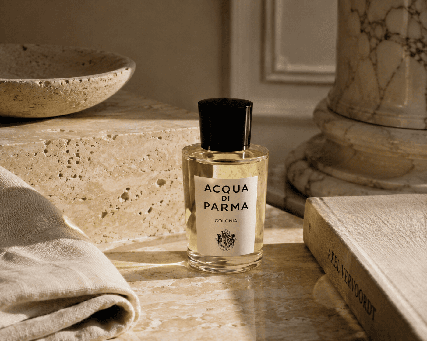 A bottle of Acqua di Parma Colonia perfume with a clear glass body, light golden liquid, cream-colored label, and a glossy black cap. The perfume stands on a warm travertine stone surface next to an Axel Vervoordt book, a travertine bowl, draped linen fabric, and a classical marble column base illuminated by soft, natural sunlight.