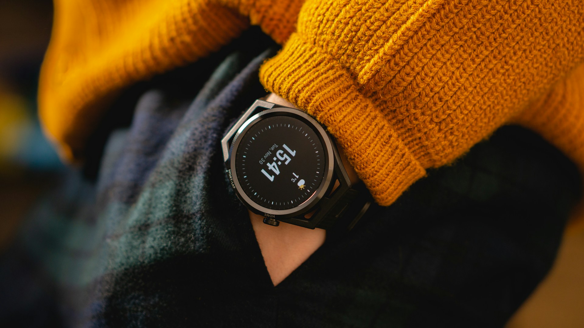 A close-up of a person's wrist wearing a dark smartwatch displaying the time 15:41 and weather information, against a background of an orange sweater and plaid pants.