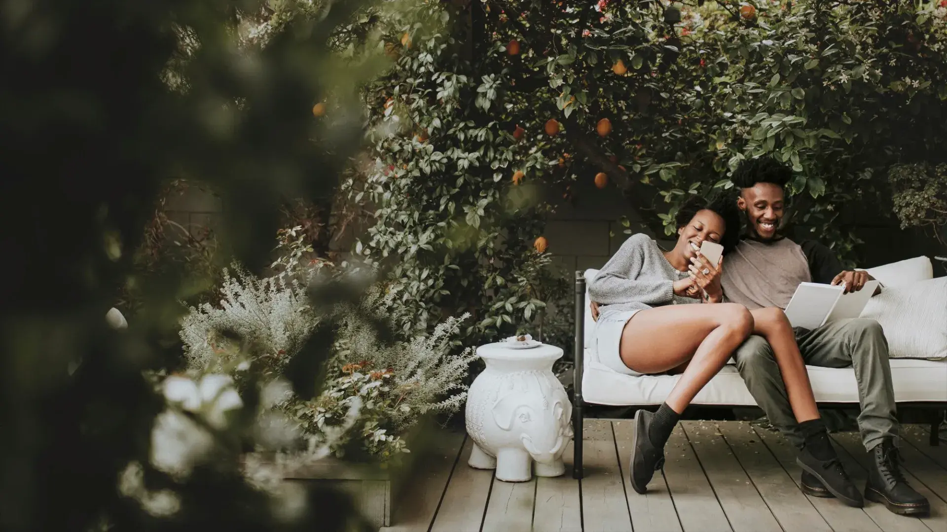 A couple relaxes together on an outdoor patio surrounded by lush greenery, smiling as they look at a phone and paperwork. The peaceful, nature-filled setting reflects the comfort and joy of homeownership through USDA Rural Development loans from Chris Lewis Home Loans.