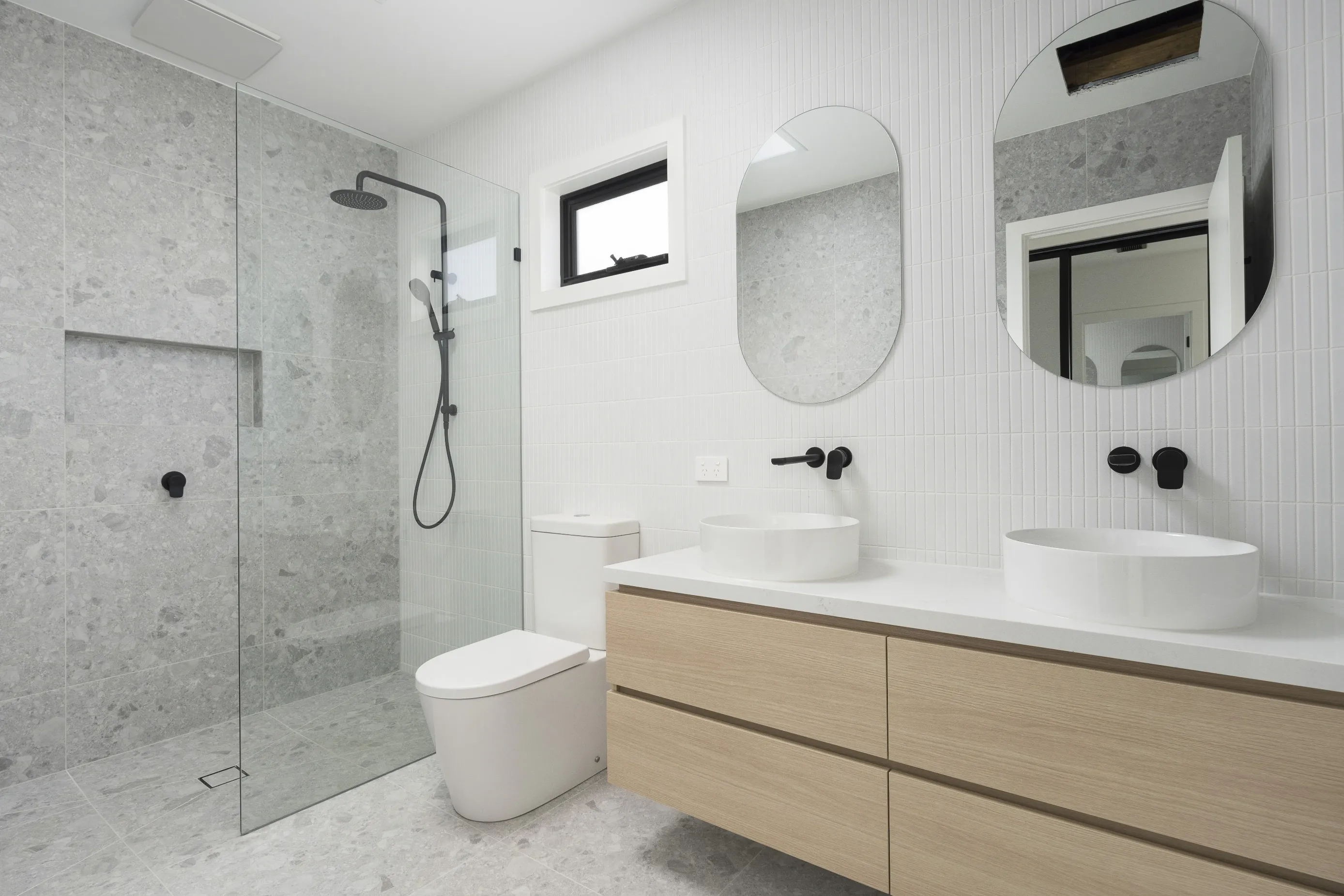 Bathroom in grey terrazzo-look tiles with a walk-in shower behind a glass screen with black rail shower, two oval mirrors, a pale-oak floating vanity with twin white vessel basins and black tapware