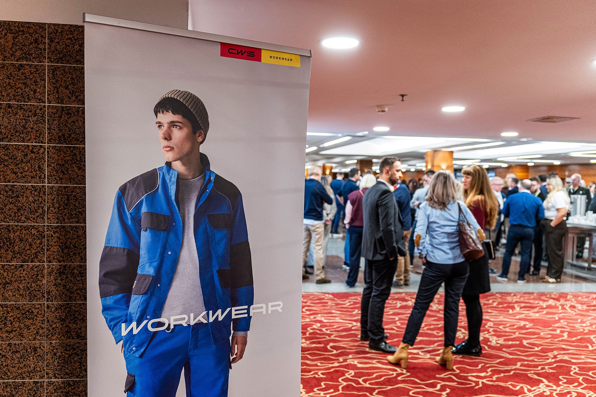 CWS Workwear banner with model in blue workwear displayed at corporate conference venue in Prague, with attendees networking in the background.