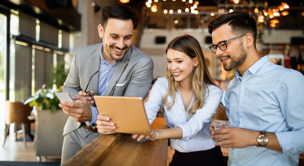 Three people, dressed in business attire, stand in a modern office environment, smiling and looking at a tablet one of them is holding. One person holds a phone and another holds a drink, discussing the benefits of revenue-based repayment for an easy loan application.