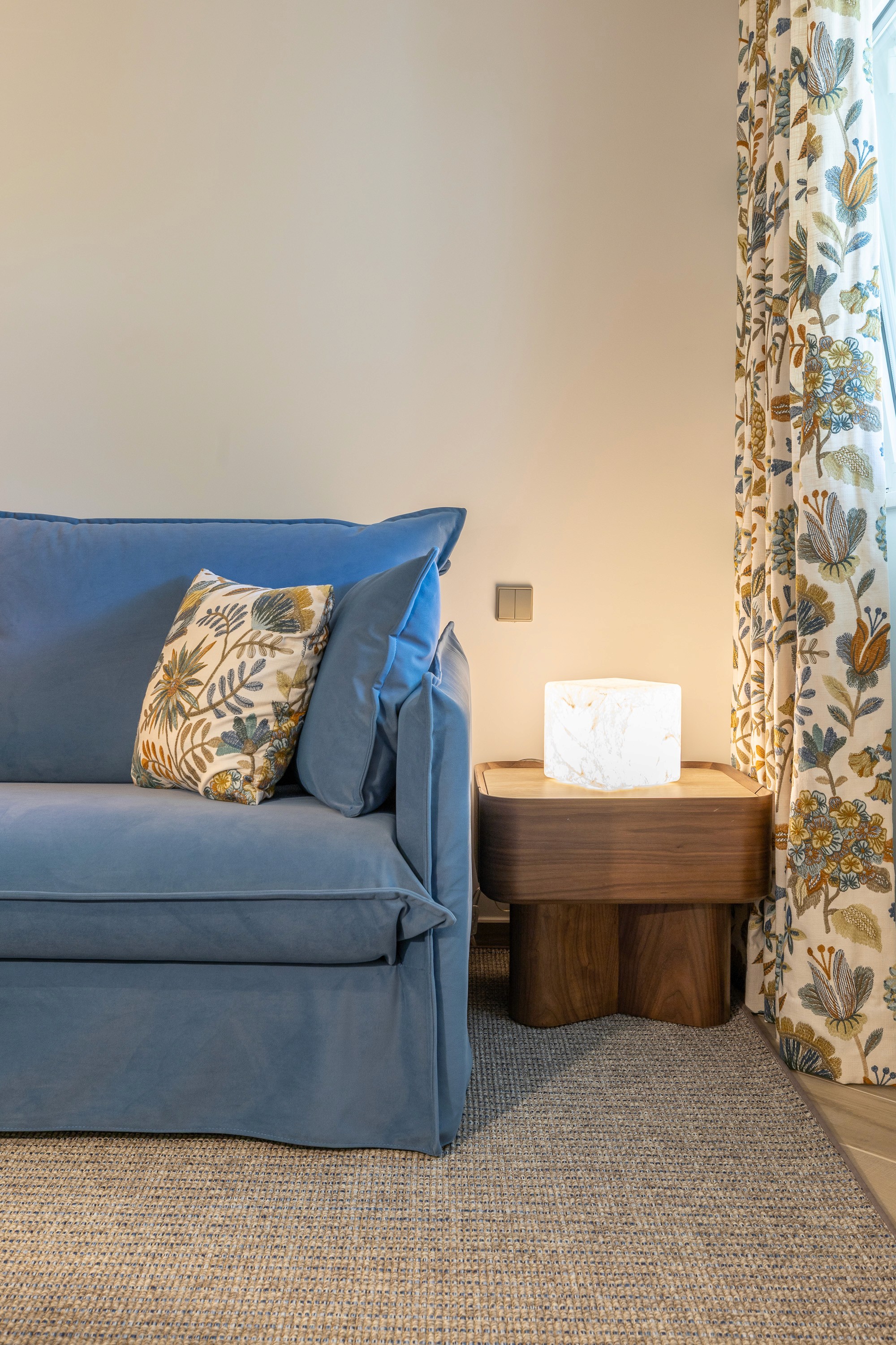 Corner of the blue sofa bed, with a wooden side table and a botanical print curtain