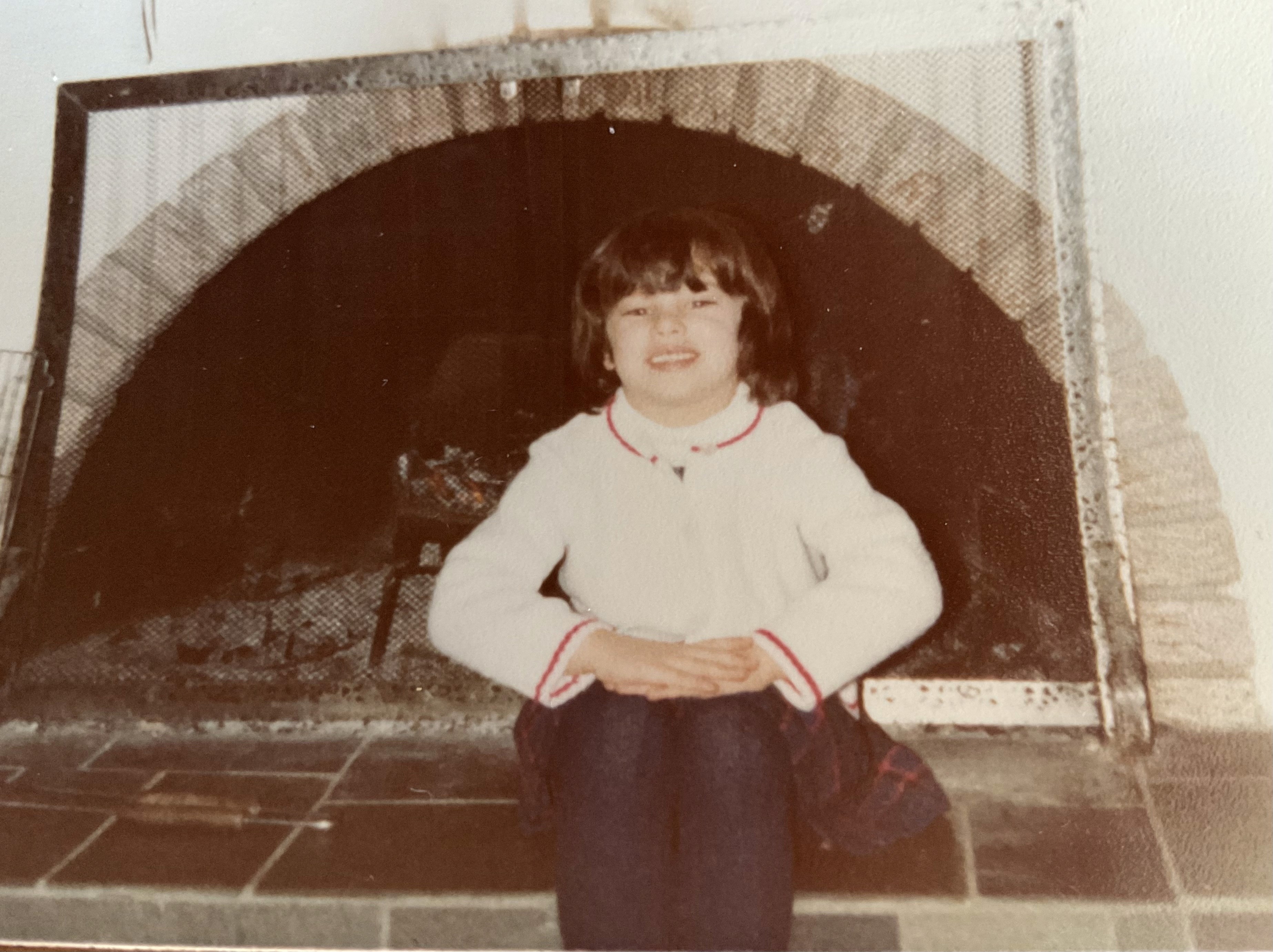 A little girl sitting on a slate hearth in front of a semi-circular fireplace wearing a white sweater and a navy skirt, December 1978. Photo by Anis Mahli.