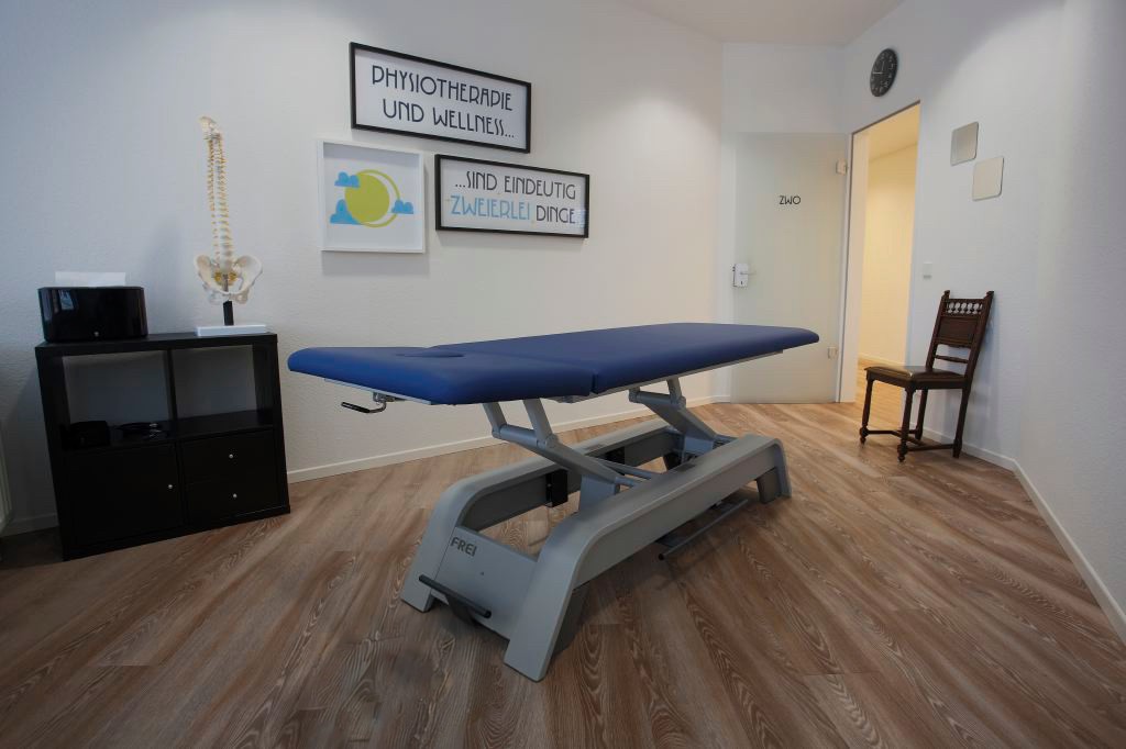 Ein Raum für Physiotherapie mit einer blauen Behandlungsbank und Stühlen, ideal für therapeutische Sitzungen und Übungen. An der Wand hängt ein Bild auf dem Text „Physiotherapie und Wellness sind eindeutig zweierlei Dinge“.