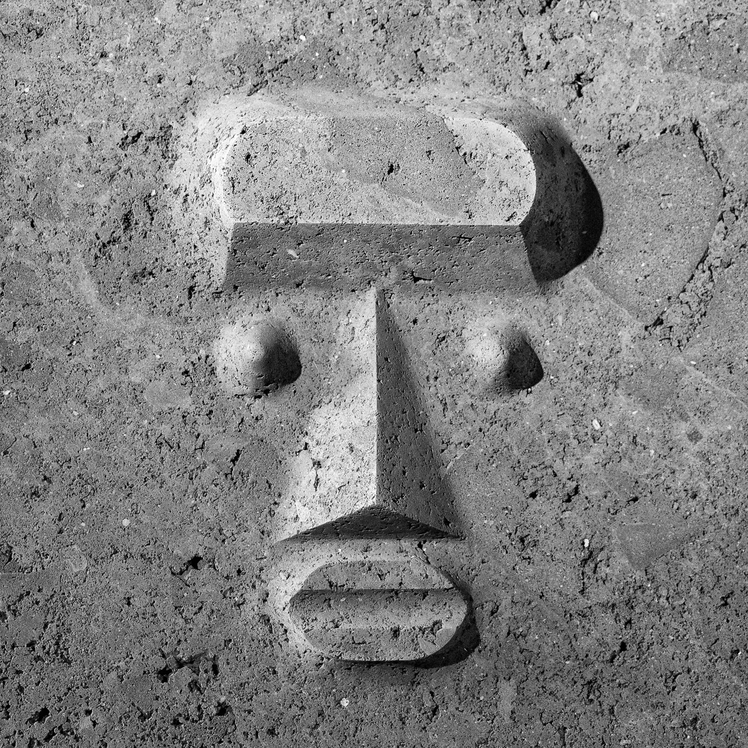 Black and white photo of a stylized face carved in volcanic stone, showing simple geometric features including eyes, nose, and lips.