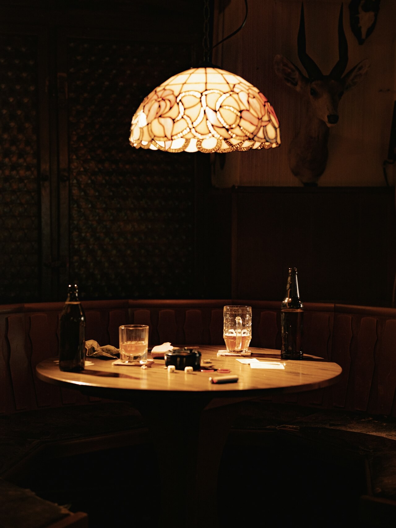Cozy, dimly lit pub scene with a round wooden table, empty beer bottles, and glasses under a stained glass lamp. A deer head decorates the wall.