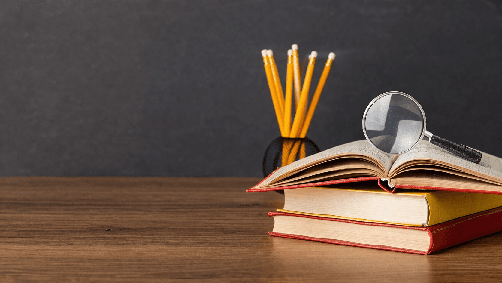 Ahşap masa üzerinde üst üste konulmuş kitapların üzerinde açık bir kitap ve büyüteç bulunan, arka planda kalemlikte sarı kalemlerin yer aldığı sade bir çalışma ortamı