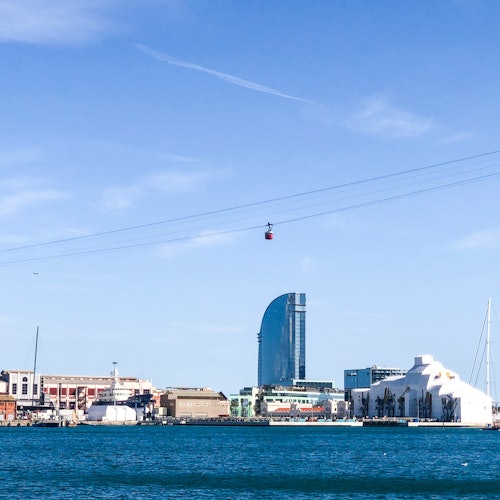 Obalni gradski krajolik s modernim neboderima, žičarom na nebu, brodovima usidrenima uz obalu i dubokim plavim morem.