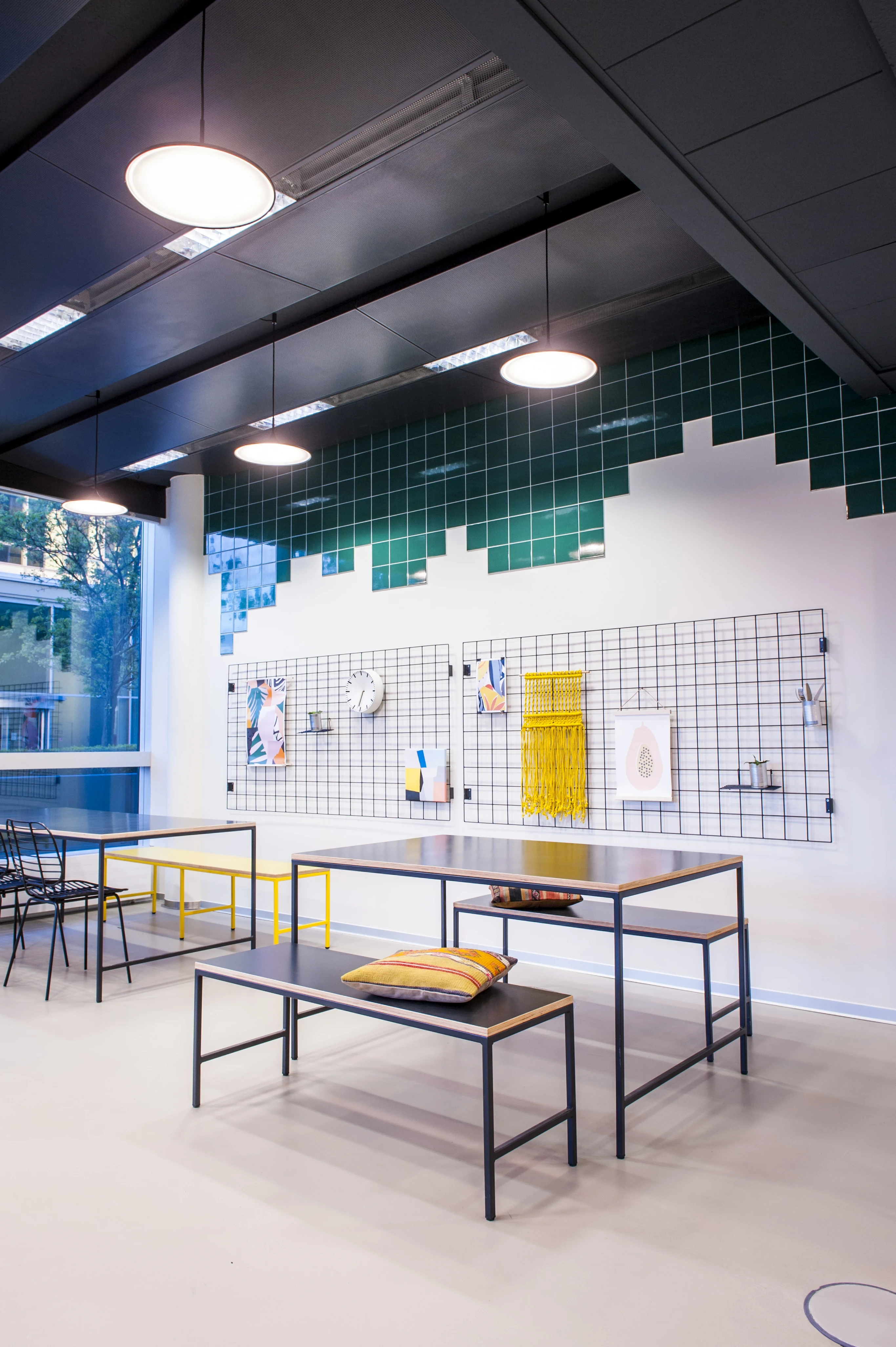 Dining area at KANTINE KOMTUR with black metal tables and benches, a yellow bench accent, wire grid wall panels with art and a pixel-style green tile pattern under a black ceiling.