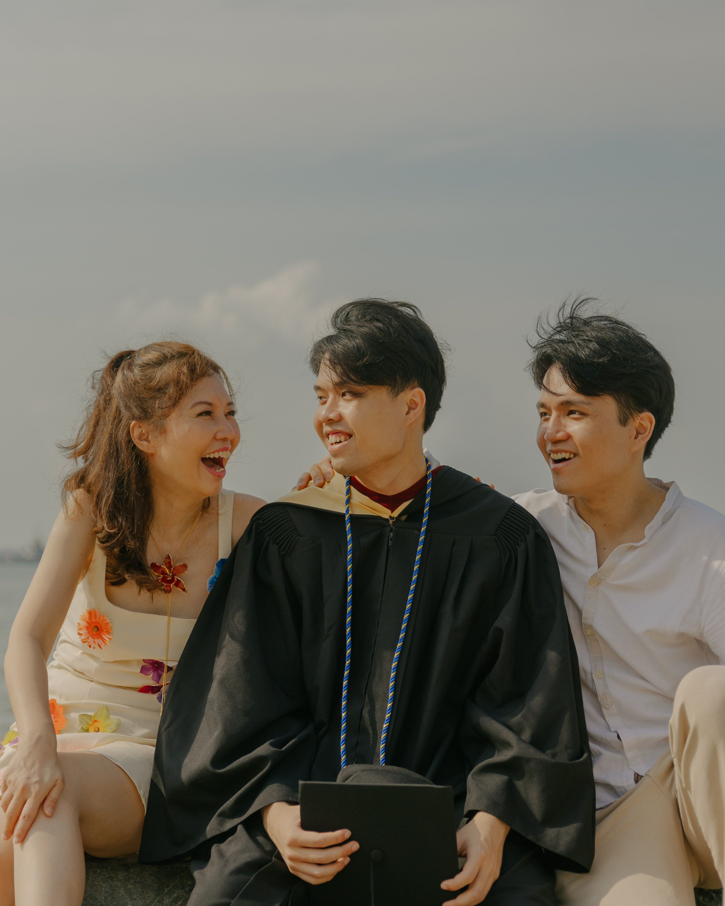 Close-up shot of the graduate and his two friends sitting and laughing together by the water's edge.