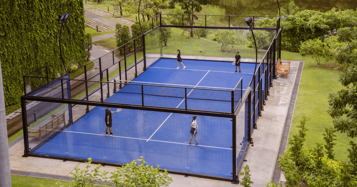 A blue outdoor padel court surrounded by greenery, with four players in a doubles match.