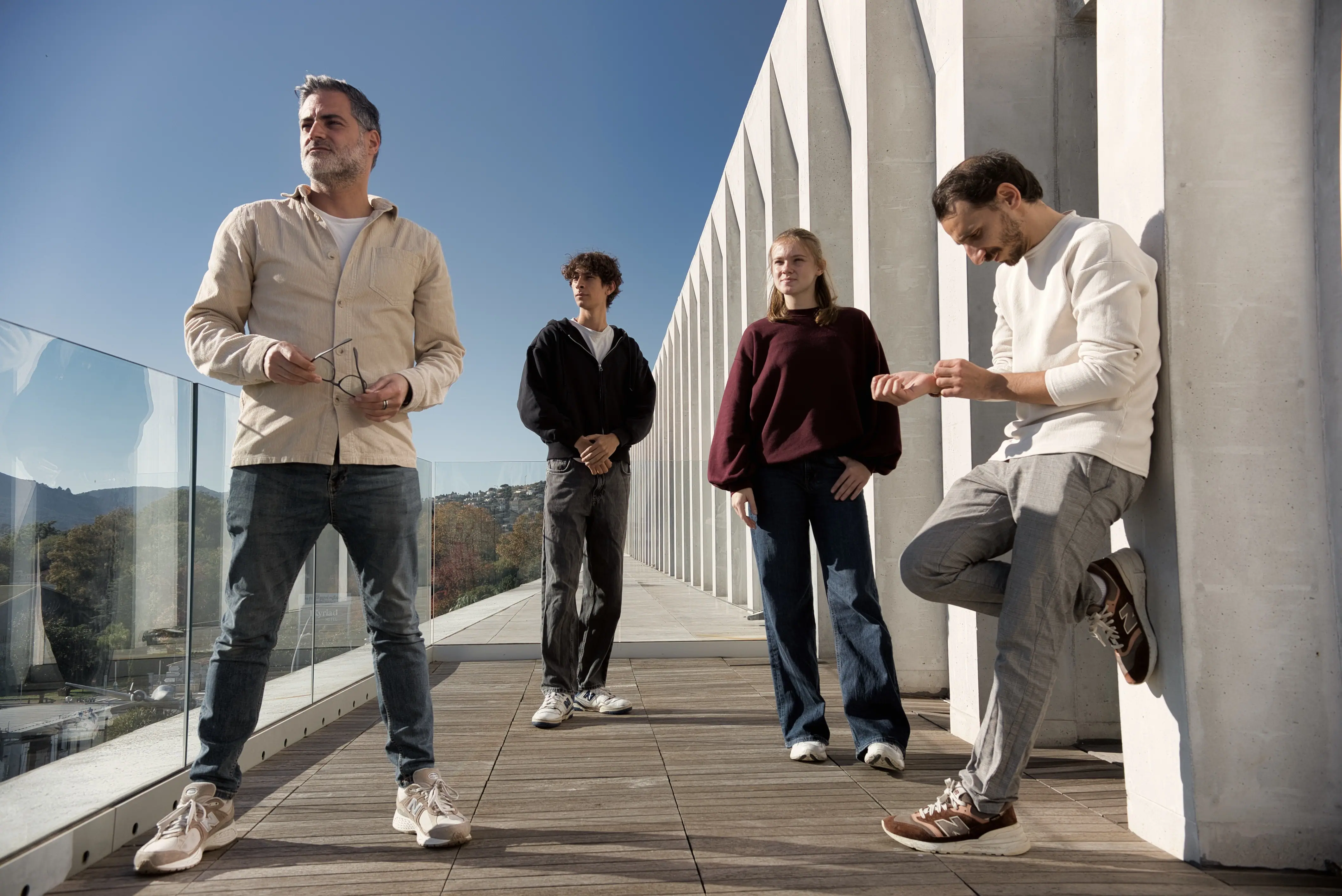 A group of four people casually standing on a modern rooftop terrace with a glass railing, featuring a clear blue sky and distant hills in the background.