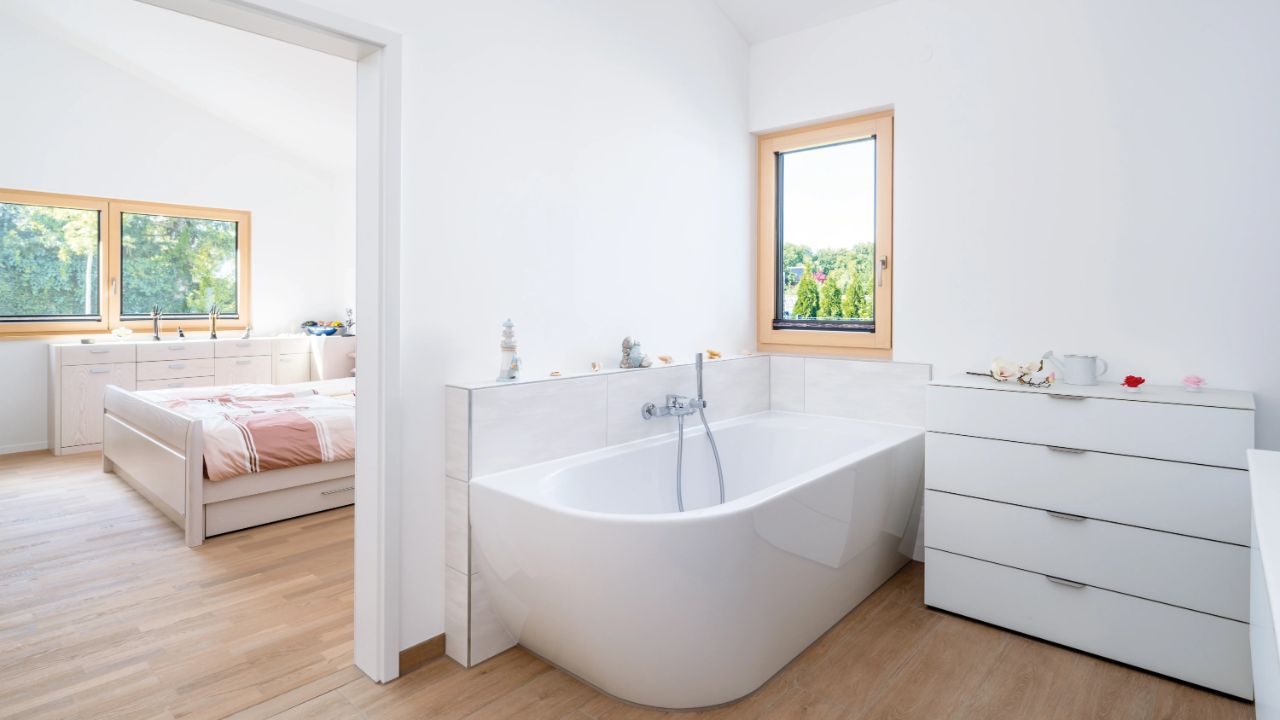 Modernes Badezimmer mit freistehender Wanne, zwei Kommoden und Blick in ein Schlafzimmer mit Bett und Fenster.