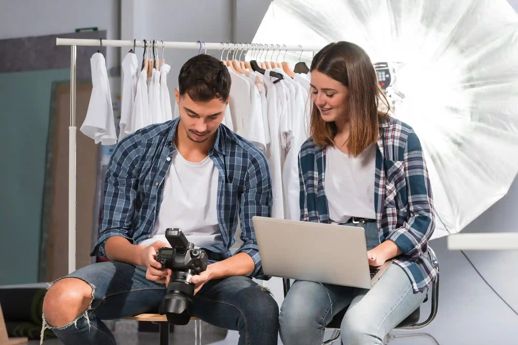 Two content creators reviewing camera equipment and laptop in a studio with clothing rack and softbox light.