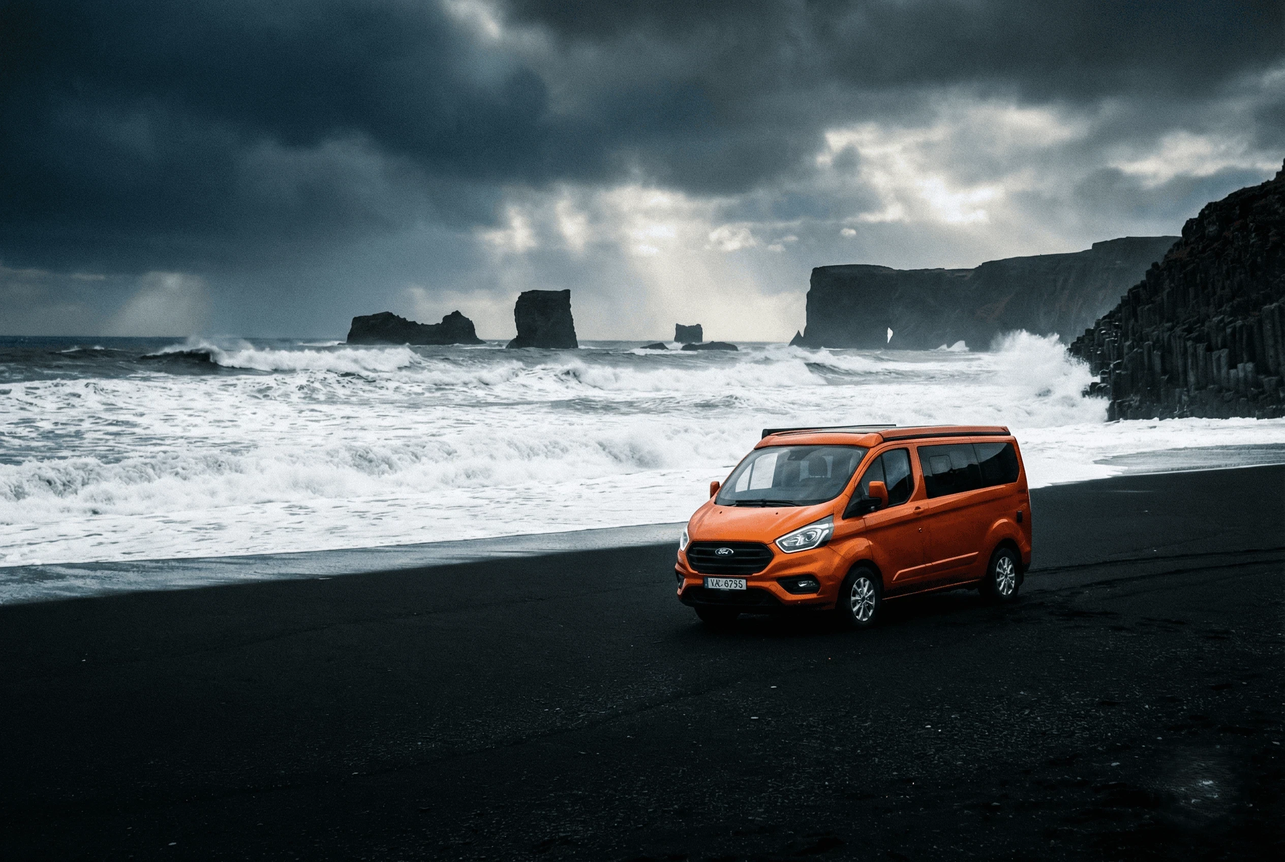 An orange camper van parked on a black sand beach with sea stacks rising from the ocean in the background.