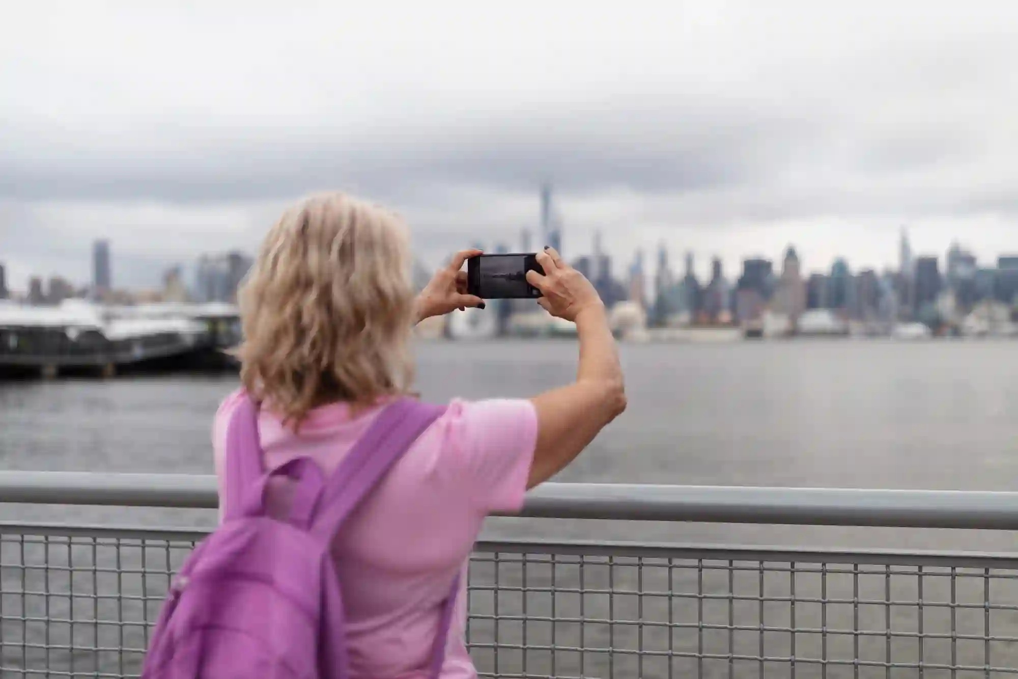 A tourist with a purple backpack photographs a city skyline across the water on a cloudy day.
