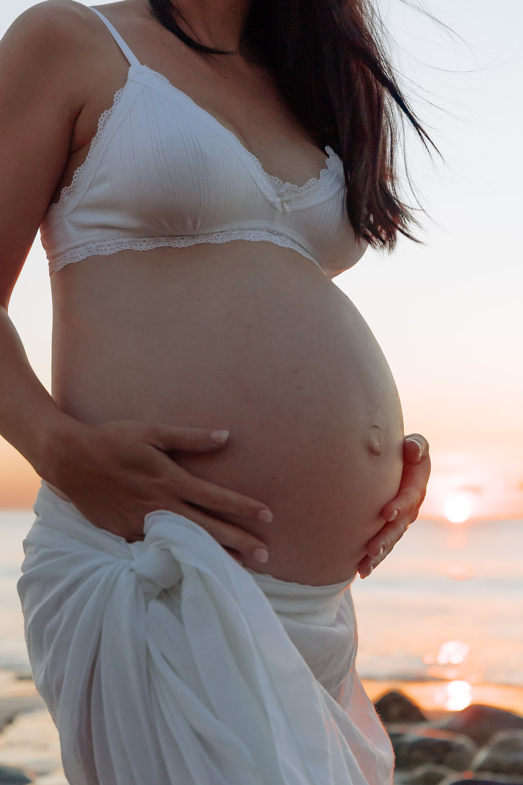Sunrise maternity portrait of a mother-to-be standing in the sand, warm morning light illuminating her profile with soft ocean tones behind her.