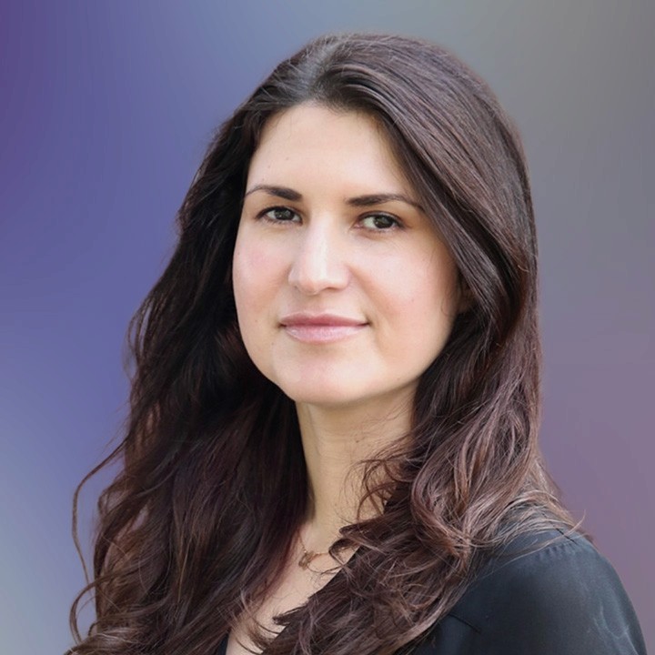 A portrait headshot of a woman with long brunette hair wearing a black blouse. with a neutral expression
