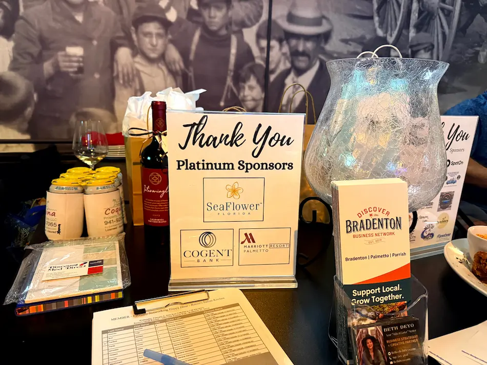 This image showcases a table display at a Discover Bradenton Business Network event, featuring sponsor appreciation signage, brochures, a glass vase, wine bottle, and beverages, underlined by the theme of supporting local businesses.