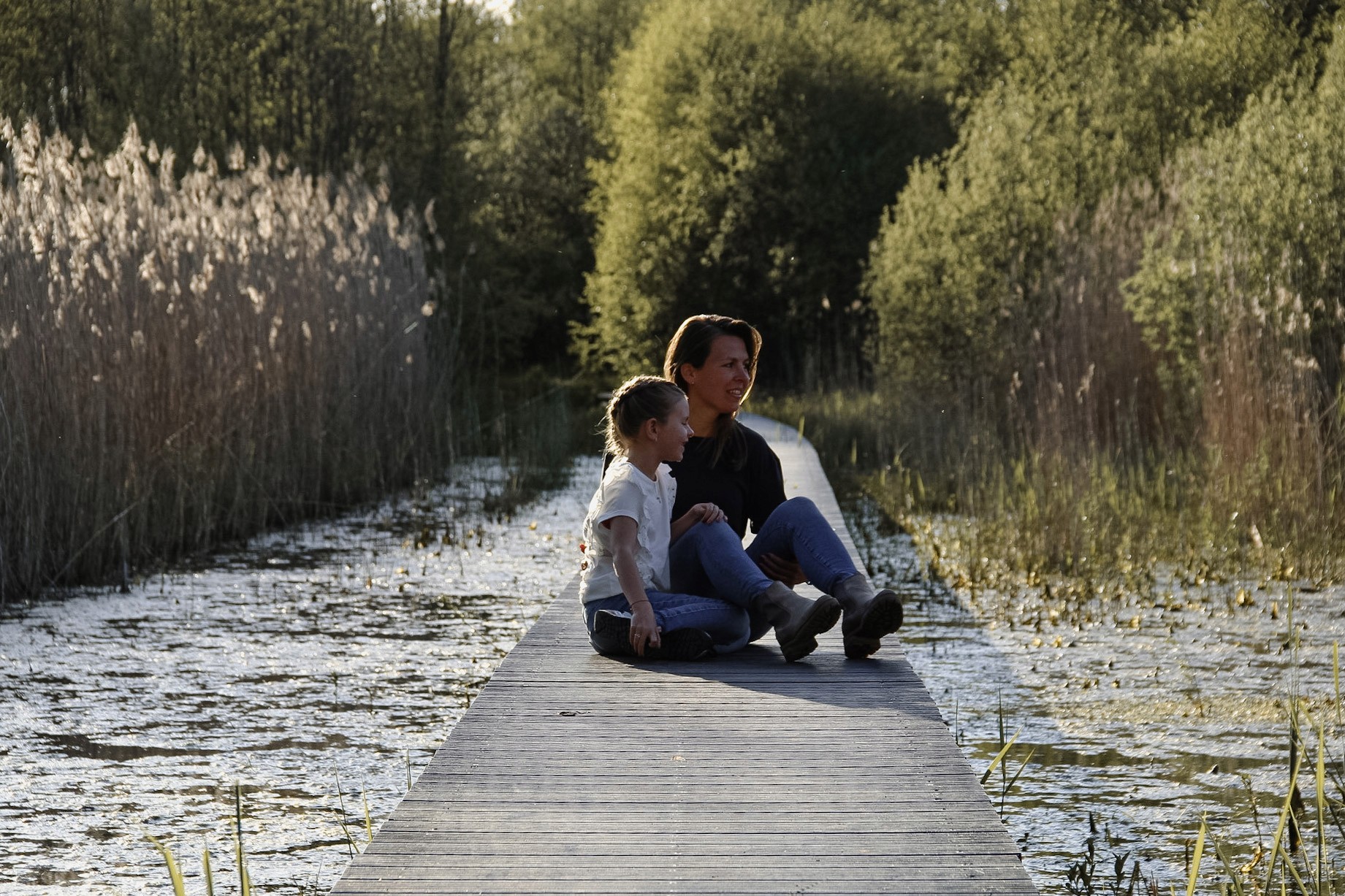 Foto van een moeder en dochter die op een kade zitten, met een focus op hun interactie en de rustige wateren op de achtergrond