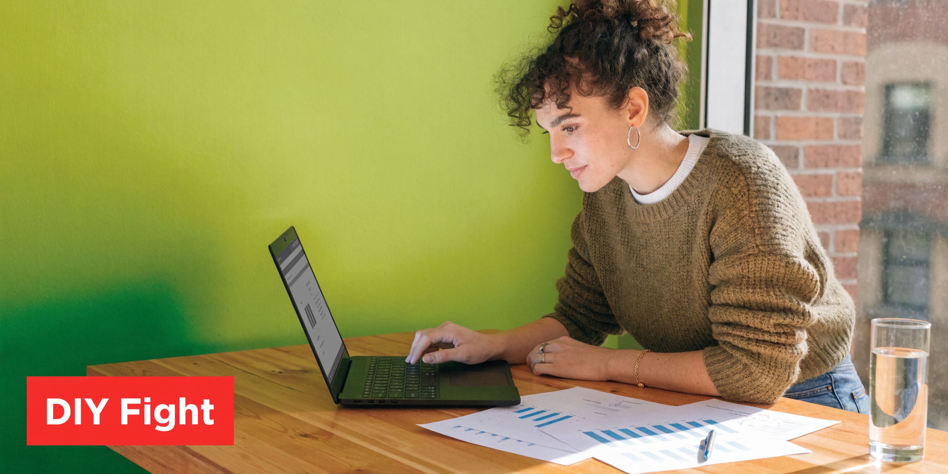 A woman working on a laptop while analyzing charts and graphs on her wooden desk at home.