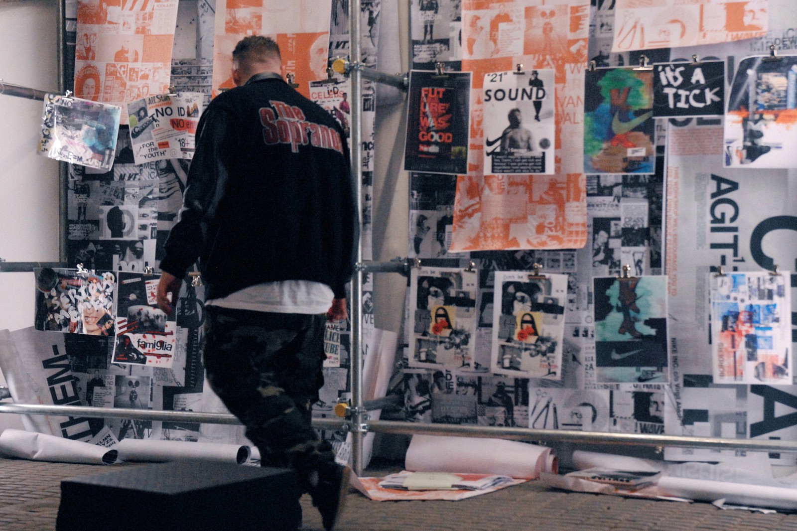 A person wearing a dark jacket is walking past a wall covered with various posters, art, and graffiti, creating an urban, creative atmosphere.