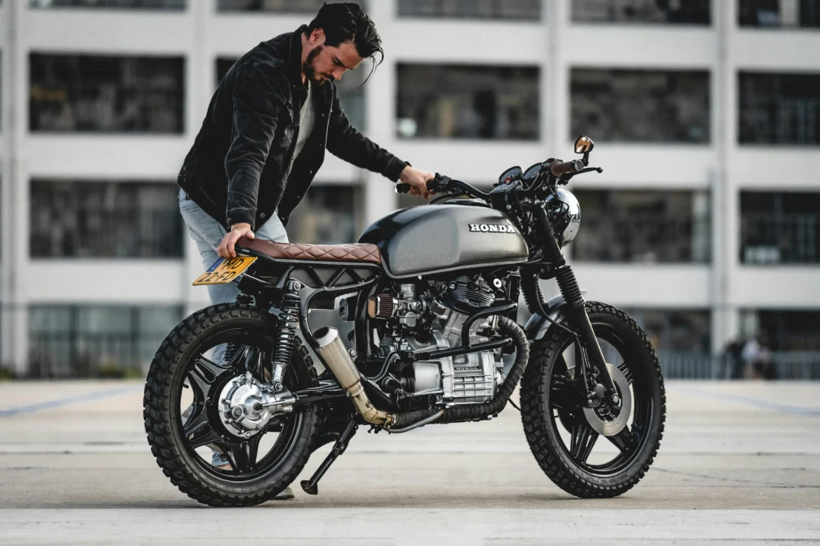 Un homme ajuste une Honda customisée, mise en valeur par un style café racer épuré. Le cadre industriel en arrière-plan contraste avec la ligne rétro de la moto, soulignant son caractère mécanique et artisanal.