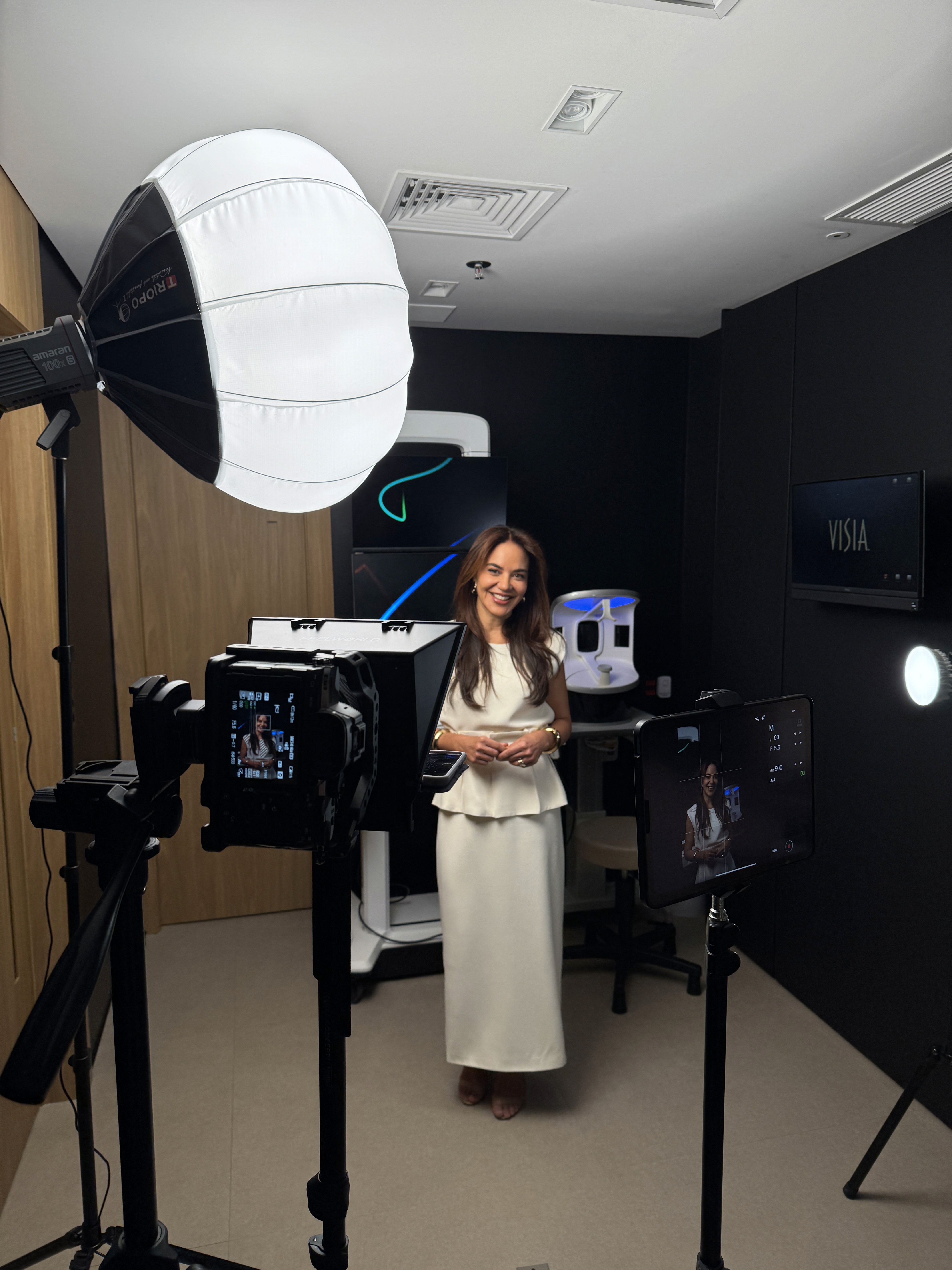 A smiling woman with her arms crossed, standing against a dark green background. She has long, dark hair.