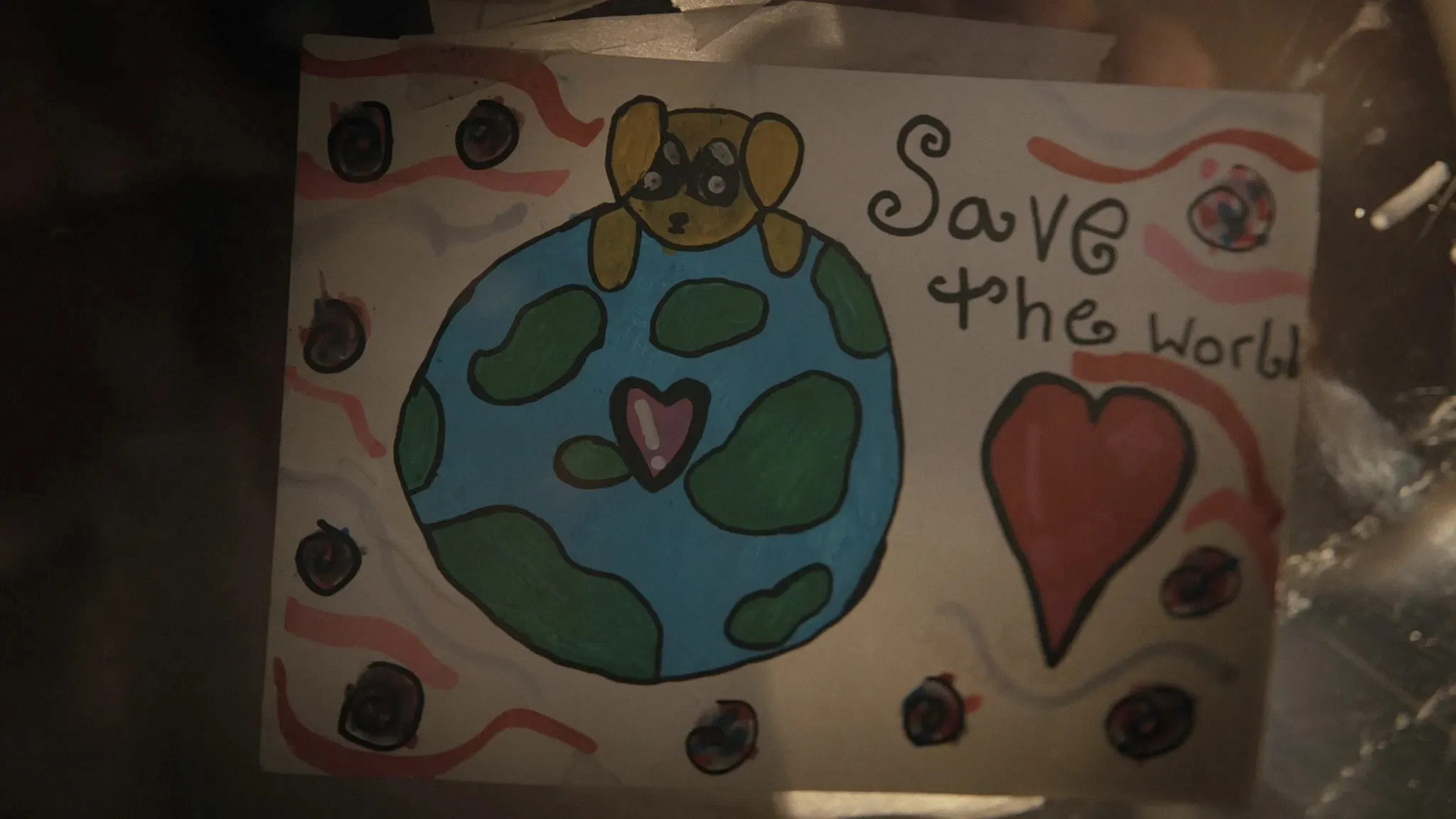 A childlike drawing depicts a cartoon dog wearing a mask sitting atop a simplified Earth, surrounded by symbols resembling planets, with the phrase "Save the world" in playful lettering alongside a large red heart.