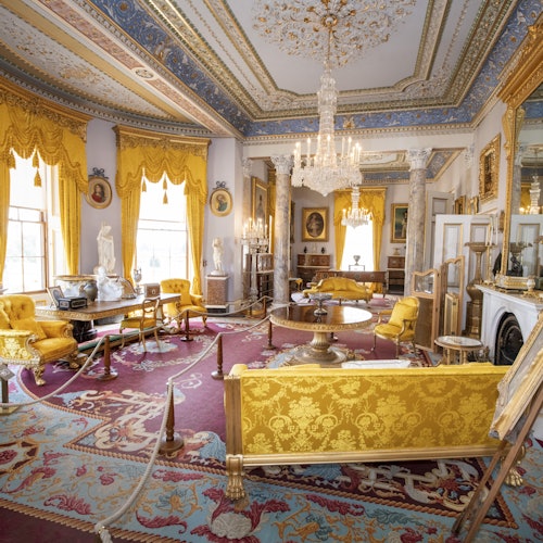 Luksuzna soba s bogatim dekorom, žutim zavjesama i namještajem, lusterima, kipovima, uokvirenim slikama i antiknim pokućstvom.