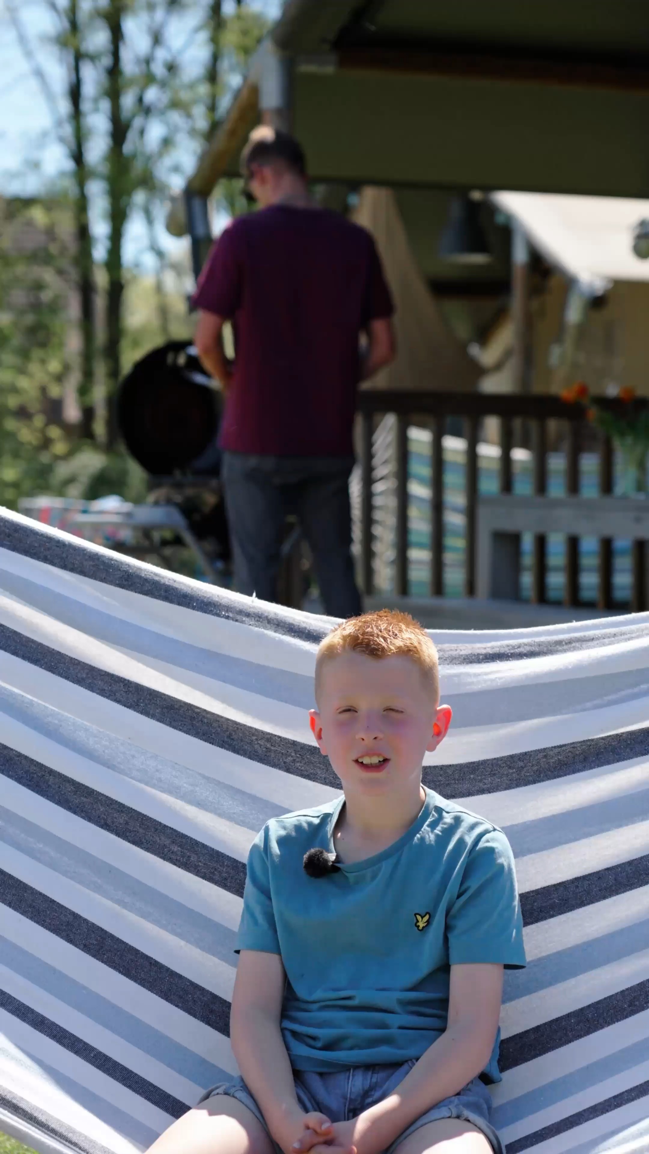 Jongen zit in een hangmat voor een safaritent, achter hem een barbecue