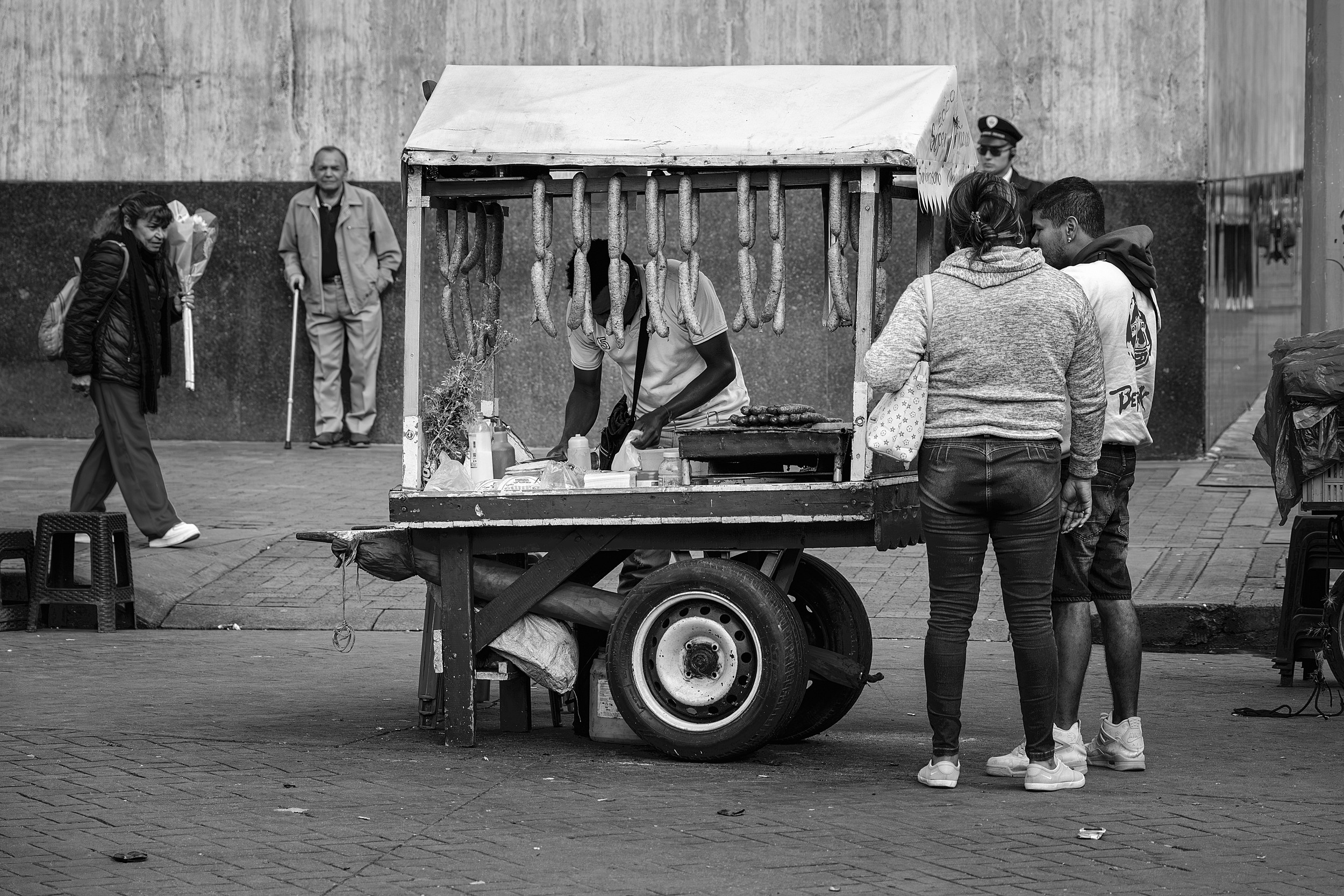 A vendor prepares grilled sausages inside a small food cart as two customers wait; strings of chorizo hang from the cart’s roof in a busy Bogotá plaza.