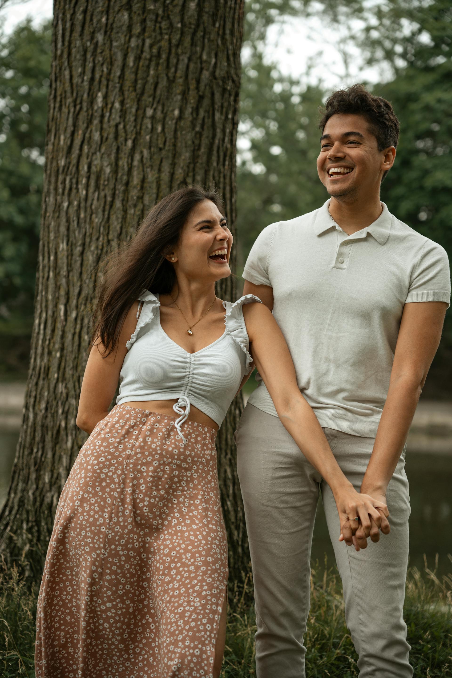 A joyful young couple laughs and holds hands while standing next to a large tree trunk in a park.