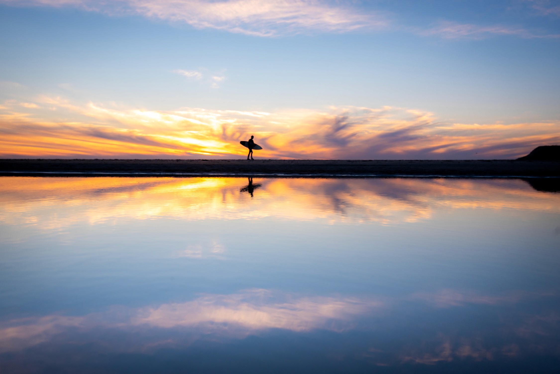 Golden sunset hues reflect across calm ocean waters, silhouetting a lone figure on the sandy shore.