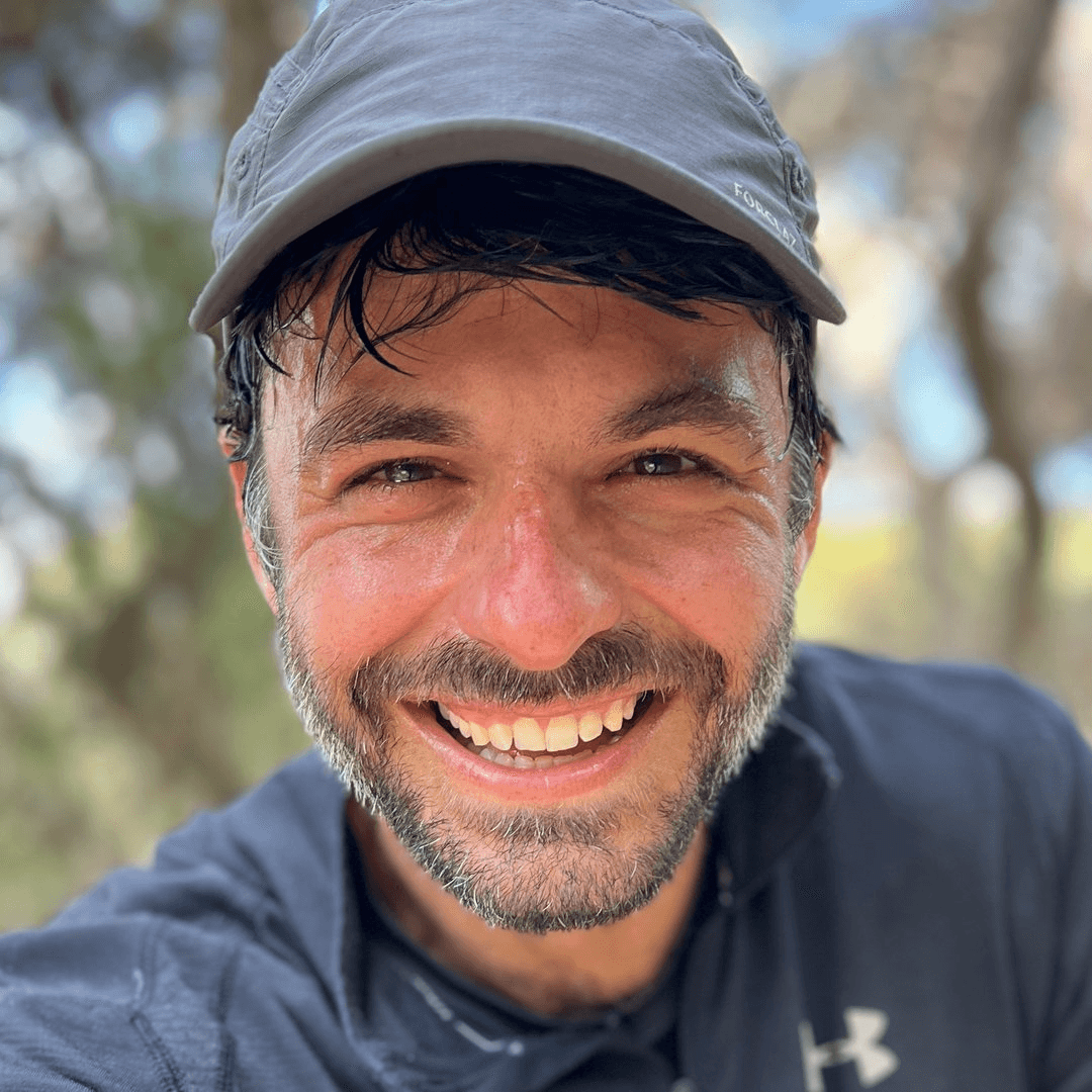 Woman in sunglasses poses against vibrant red background, wearing a beige blazer and black top. Sunlight casts dynamic shadows, creating a stylish, confident mood.
