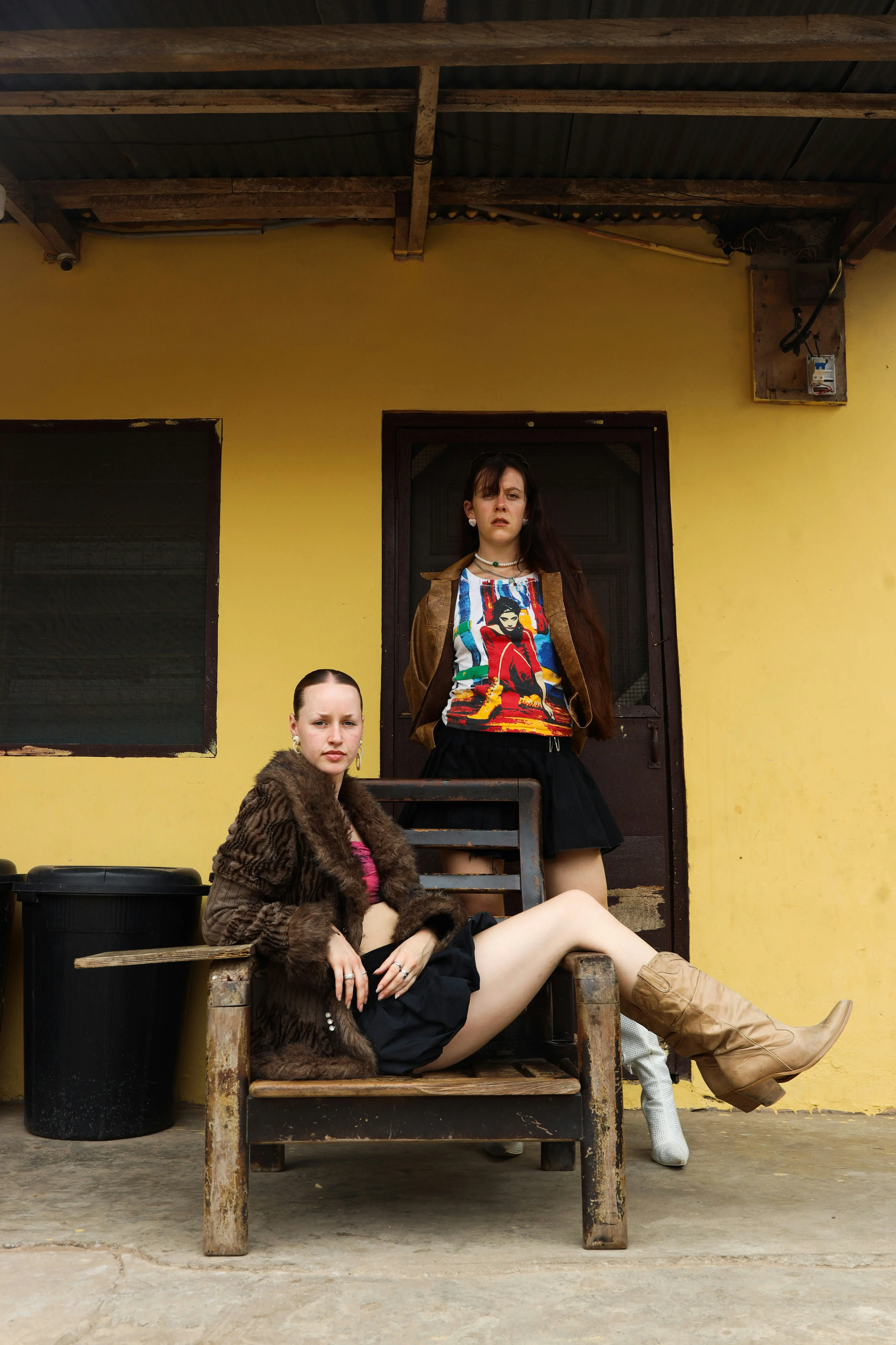 Two stylish women posing in an outdoor urban setting against a mustard yellow wall, showcasing a mix of faux fur, graphic prints, and modern layered fashion.
