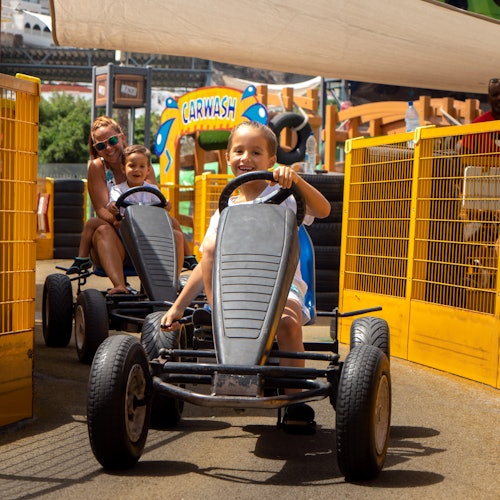 在一个阳光明媚的日子里，两个孩子在成人的监督下驾驶卡丁车，背景中有一个“洗车”的标志。
