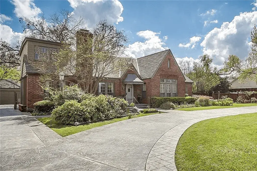 Large brick suburban home with a curved driveway and landscaped front yard under a partly cloudy sky.