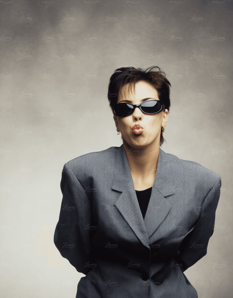 Person in oversized gray blazer and sunglasses puckering lips playfully against a neutral gray background.