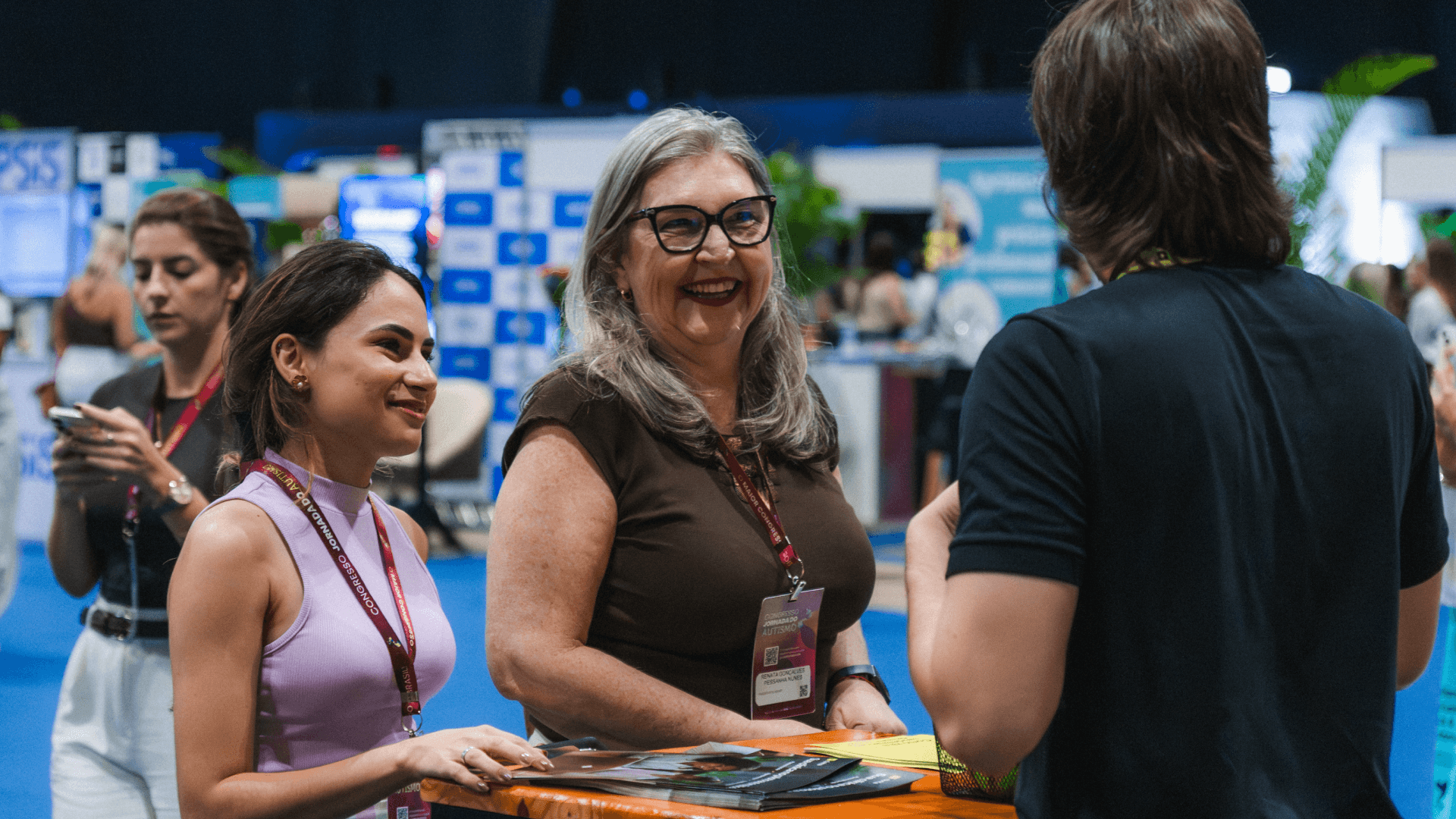 Representante da Tivita sorrindo durante atendimento no estande do Congresso Jornada do Autismo, com materiais institucionais sobre a mesa, em ação de relacionamento com profissionais de clínicas especializadas no Rio de Janeiro