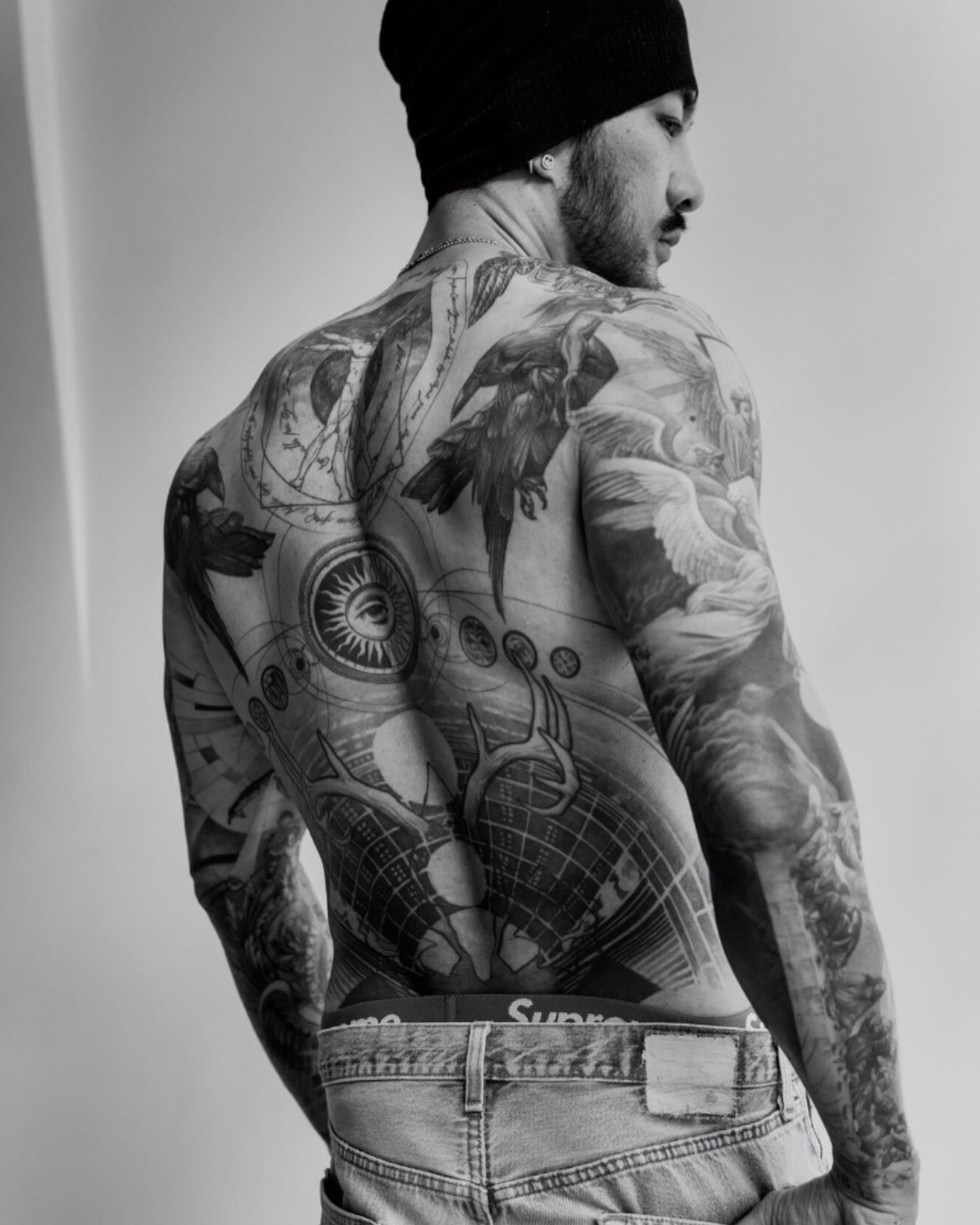 A man with a full back tattoo featuring detailed, symmetrical designs that include eyes, birds, and geometric shapes.