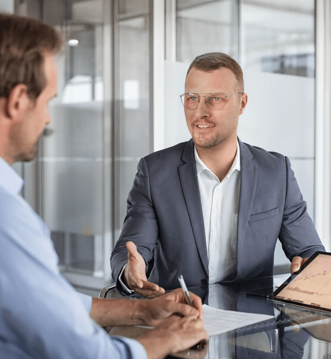 Zwei Geschäftspartner in einem Besprechungsraum; ein Mann mit Brille im Anzug führt ein Gespräch, während sein Gegenüber sich Notizen macht und ein Tablet auf dem Tisch liegt.