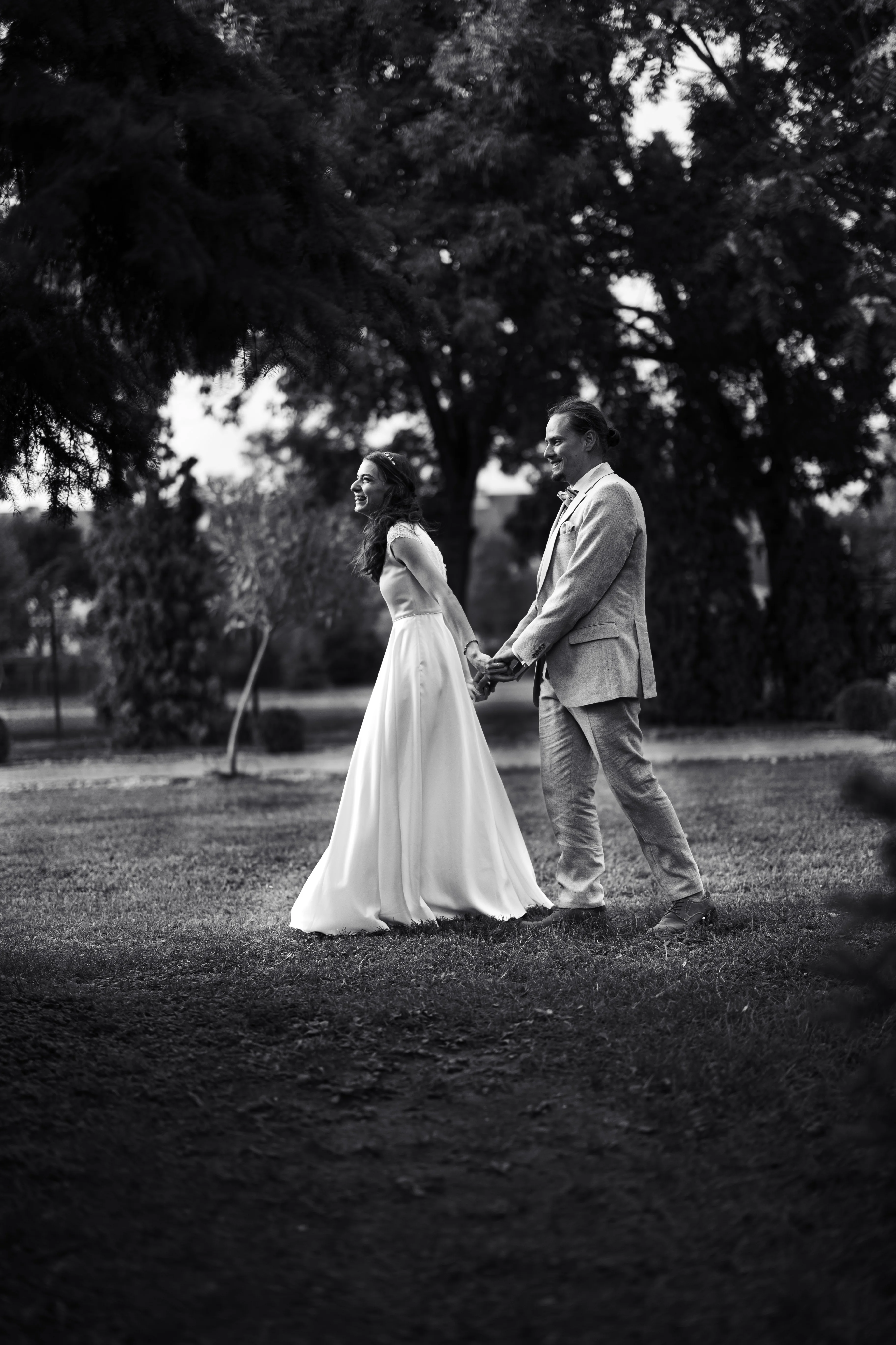 Fekete-fehér esküvői fotó: menyasszony hosszú fehér ruhában és vőlegény bézs öltönyben kézen fogva állnak füves parkban fák között, Hatvan
