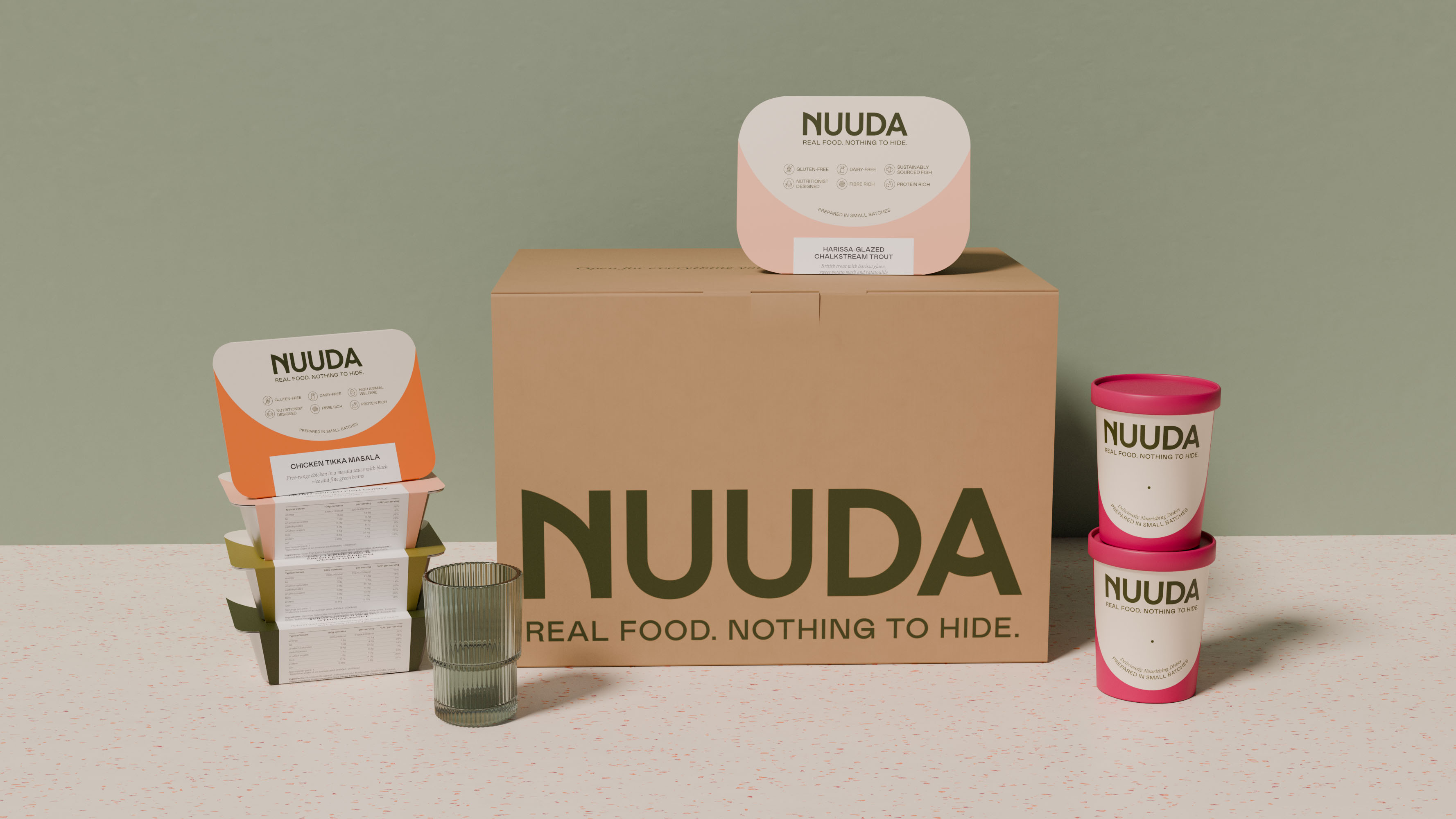 Full Nuuda product range displayed together: a large delivery box, stacked meal sleeves in varying colours with nutritional label visible, and two meal pots, showing the complete packaging system