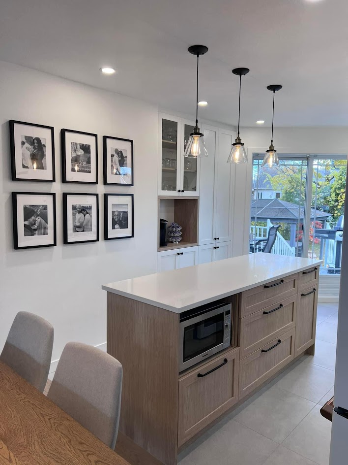 Cuisine blanche avec îlot en bois blond pâle et un comptoir de quartz blanc. Un mur décoratif présente six cadres aux contours noirs. L,espace café, entouré de bois, rappelle le matériaux de l'îlot et crée une belle continuité visuelle. Une porte vitrée s'ouvre sur la cuisine, complétée par des poignées noires et des armoires de style shaker. Dans le coin gauche, une table en bois avec deux chaises en tissu vient finaliser l'aménagement.