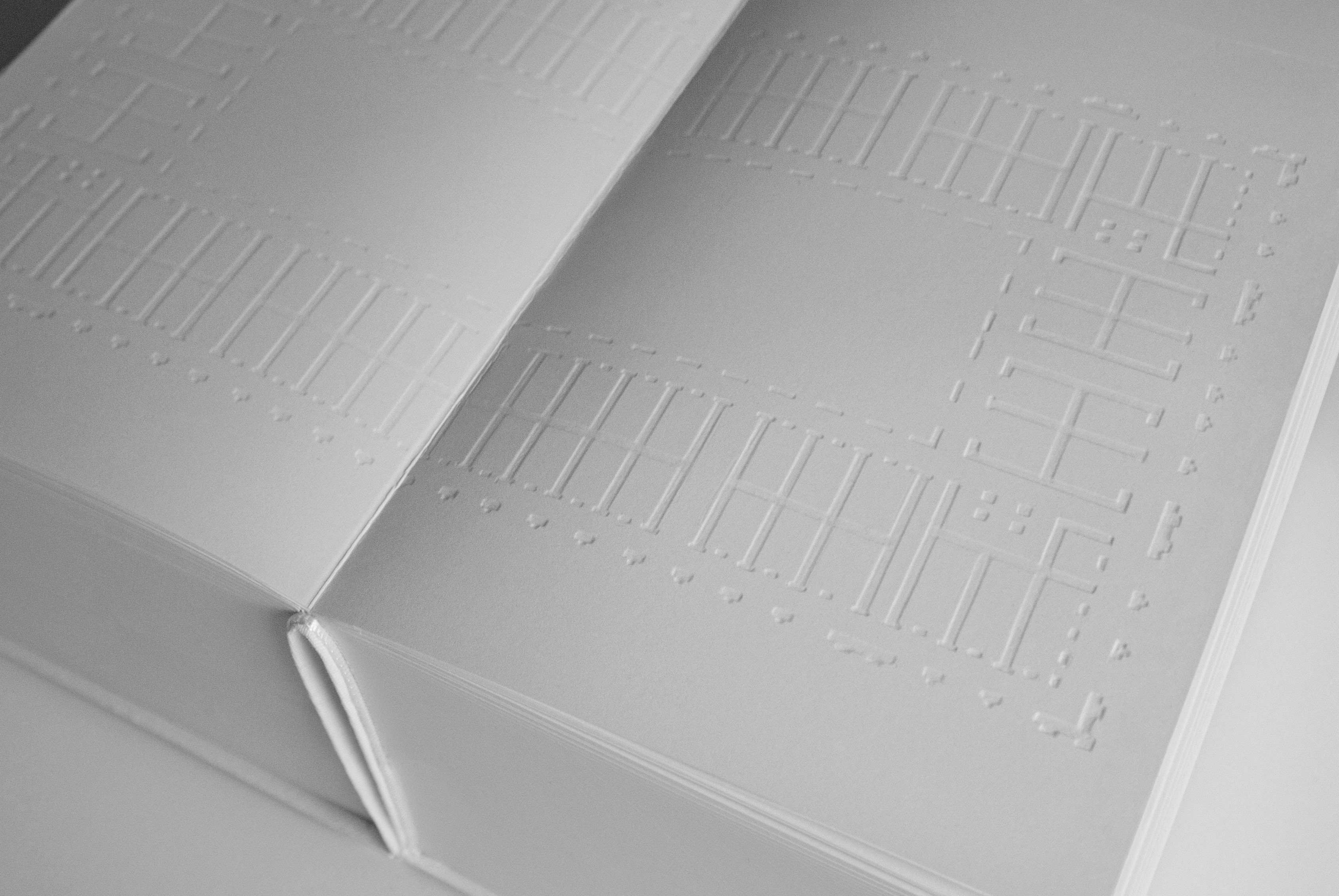 Close-up of an open white book with embossed architectural lines and window-like grid patterns pressed into the pages, creating a tactile floor plan visible only through light and shadow.