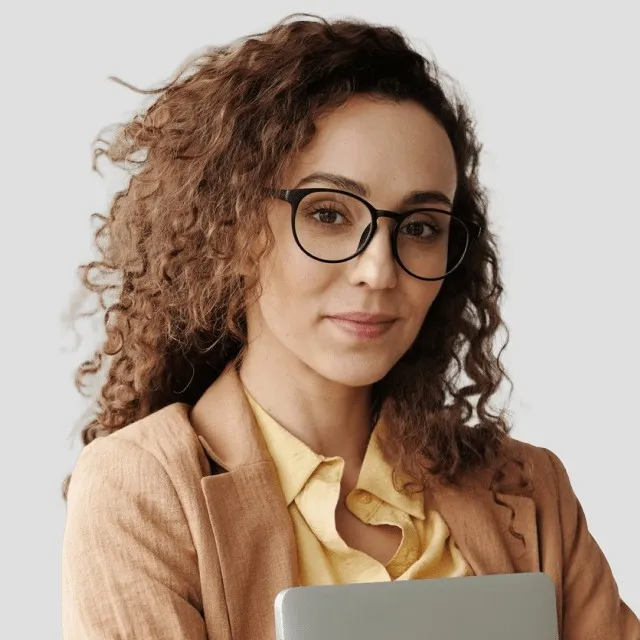Woman with curly hair and glasses holding a laptop on a neutral background smiling slightly.